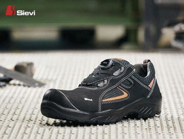 The image shows a black work shoe with a round toe cap. It has orange accents and a non-slip sole. The shoe is placed on a gray, textured surface.