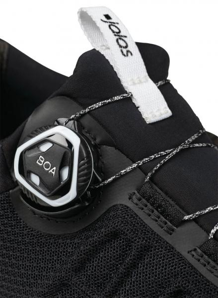 Das Bild zeigt einen Teil eines schwarzen Schuhs mit einem drehbaren Verschlusssystem. Der Schuh hat eine strukturiert Oberfläche und einen weißen Schriftzug oben.