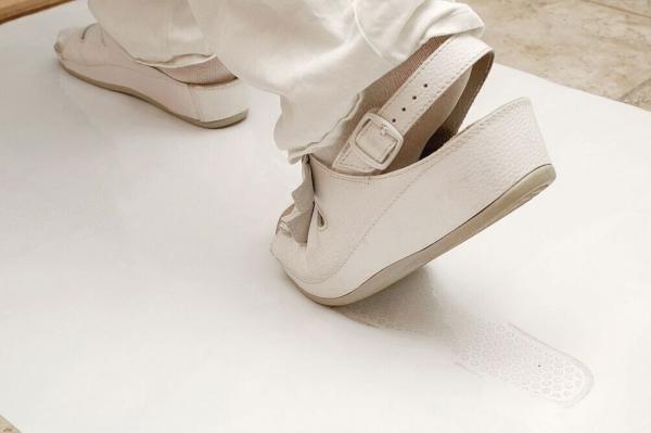The image shows a foot in white shoes on a smooth, light floor. The shoe has an adjustable strap. The background is blurry, and the focus is on the shoe.