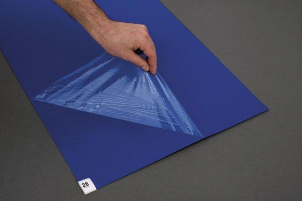 A hand lifts a transparent, wavy film lying over a blue area. The film has a triangular pattern and reflects the light. In the background is a gray surface.
