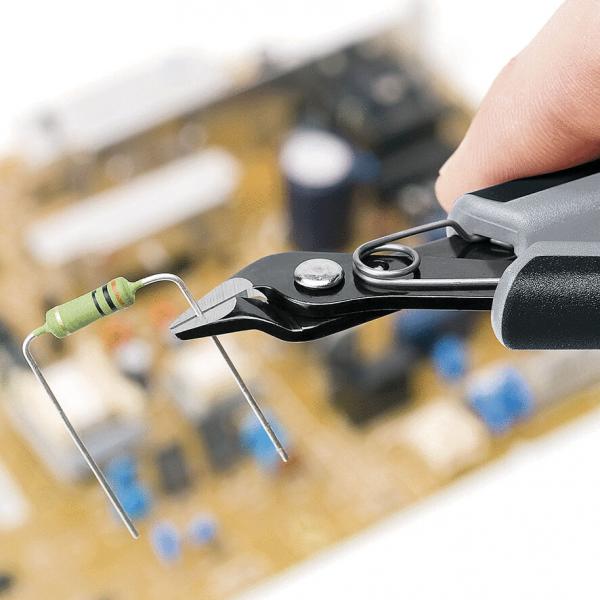 In the picture, you can see a hand holding a pair of pliers that is gripping a resistor. In the background, a circuit board with various electronic components can be seen out of focus.