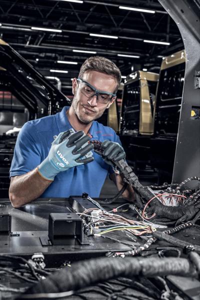 A young man is wearing safety goggles and gloves while working on a vehicle. He is checking cable connections in a busy workshop environment.
