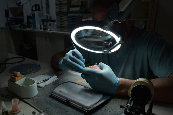 A person is wearing gloves and working under a magnifying lamp. In front of them lies a processed model. The room is dimly lit to enable precise work.