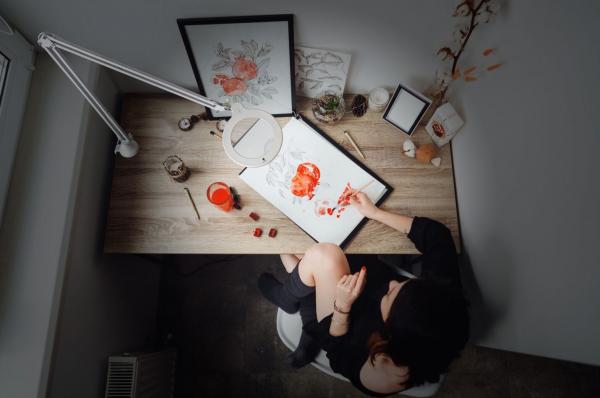 The image shows a table with art supplies. A person is sitting there, painting with reddish colors. Around the table, there are several objects, including a frame and a plant.