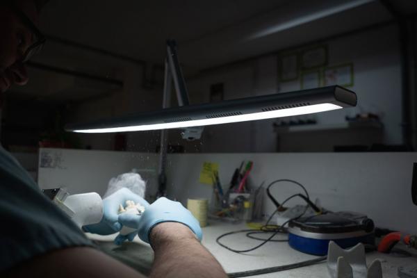 Auf dem Bild sieht man einen Arbeitstisch mit einer hellen Lampe. Eine Hand mit blauen Handschuhen hält etwas Kleines. Um den Tisch liegen verschiedene Utensilien und Materialien.