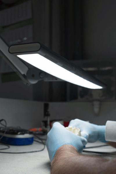Ein Tisch mit einer hellen, schwenkbaren Lampe. Eine Hand in blauen Handschuhen hält ein kleines Objekt. Im Hintergrund sind unscharfe Werkzeuge und Geräte.