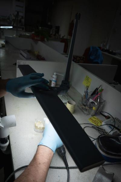 Auf dem Bild sieht man eine Hand mit blauen Handschuhen, die eine schwarze Lampe über einem hellen Tisch greift. Im Hintergrund sind verschiedene Objekte und Schreibtischutensilien sichtbar.