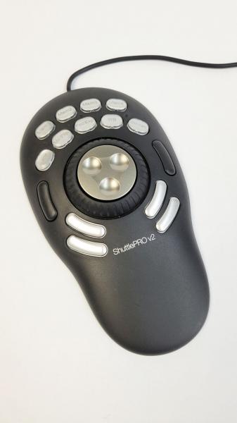 Das Bild zeigt einen ovalen, schwarzen Controller mit einer zentralen runden Drehknopf und mehreren Tasten. Der Controller hat eine kabelgebundene Verbindung und ist für multimediale Anwendungen gedacht.