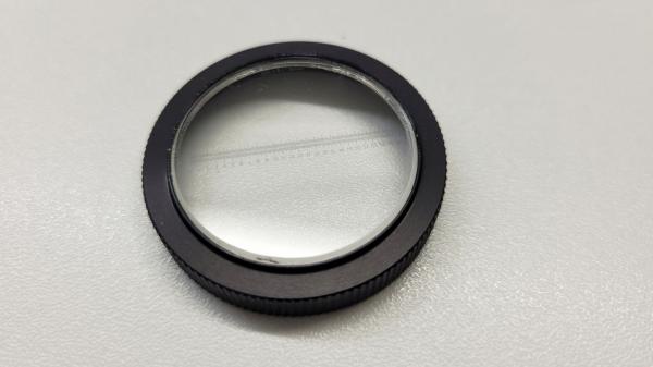 The image shows a round, flat lens frame made of black metal. In the center, there is a clear, transparent lens. The edge has a ridged structure for better grip.