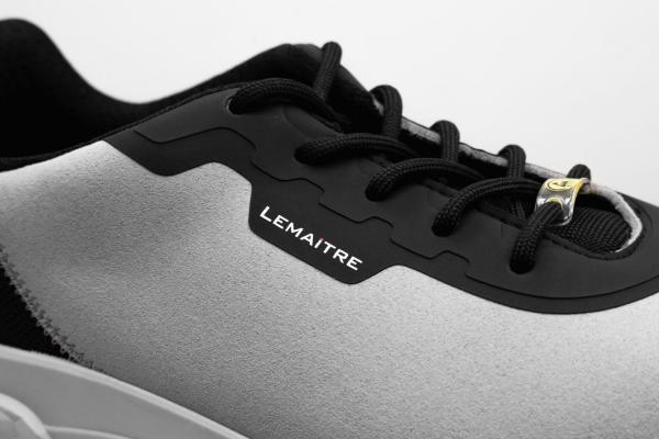 Das Bild zeigt die Nahaufnahme eines Sportschuhs. Der Schuh hat eine graue Oberseite mit schwarzen Details und Schnürsenkeln. An der Seite befindet sich das Logo ?Lemaitre?.