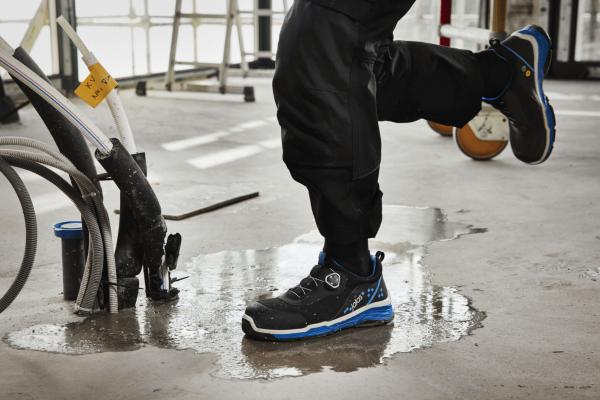 A foot stands on a wet floor, surrounded by tools and hoses. The shoe is black with blue accents, the pants are dark and protect against moisture.