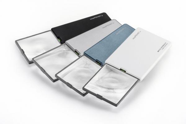 The image displays several flat magnifying lenses in various colors: black, silver, blue, and white. They are arranged side by side, some are open and reveal the lens.