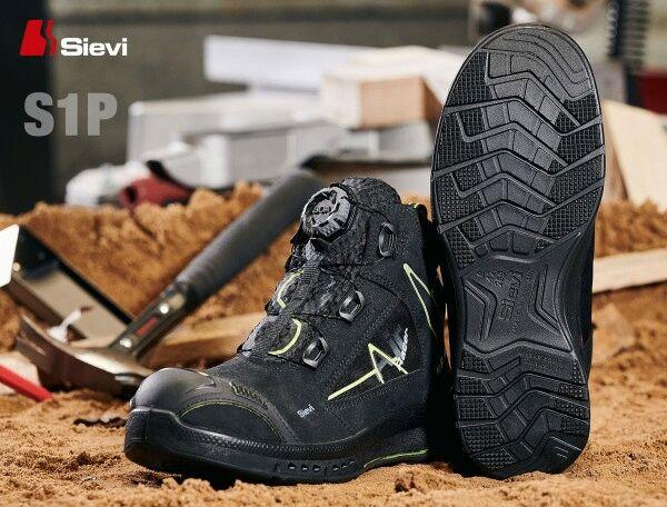 The image shows a black safety shoe with yellow accents and a sturdy sole. The shoe features a quick-lacing system and is placed on a sandy surface, surrounded by tools.