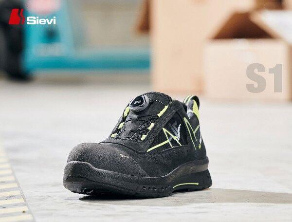 The image shows a black safety sneaker with yellow accents. It features a sturdy, reinforced toe and a quick lacing system. The shoe is on a gray floor, surrounded by packaging.