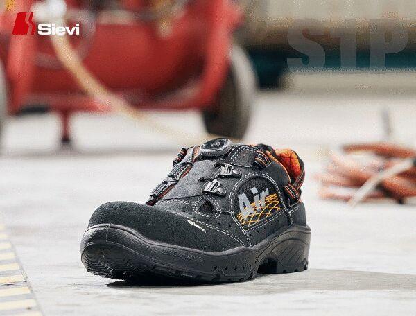 The image shows a safety shoe on a concrete floor. The shoe is black with orange accents and has a sturdy, non-slip sole. In the background, cables and a blurred machine element can be seen.