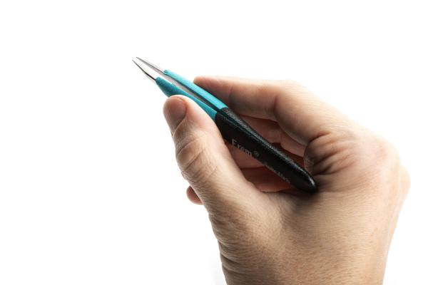 A hand is holding a blue pen with a black grip area. The pen has a pointed tip pointing upwards. The background is white and neutral.