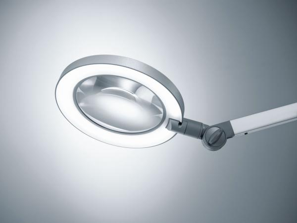 The image displays a magnifying lamp with a round, transparent glass and an integrated ring of light. The arm is flexible and allows for various positions.