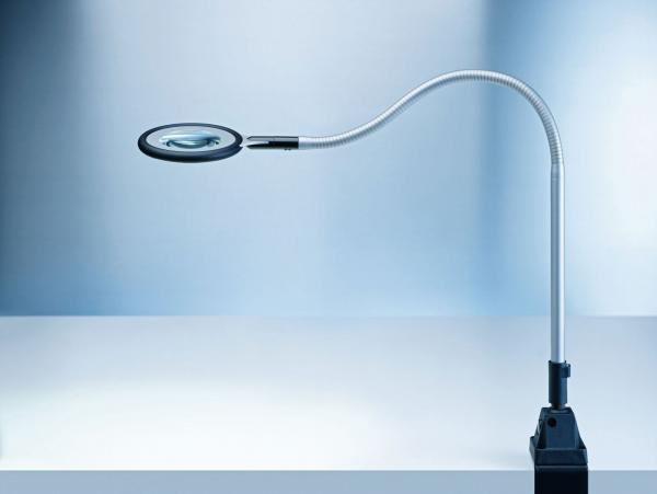 The image displays a flexible magnifying glass with a round lens at the end of a curved arm. The arm is mounted on a sturdy, rectangular base that supports the lens. The background is bright and monochromatic.