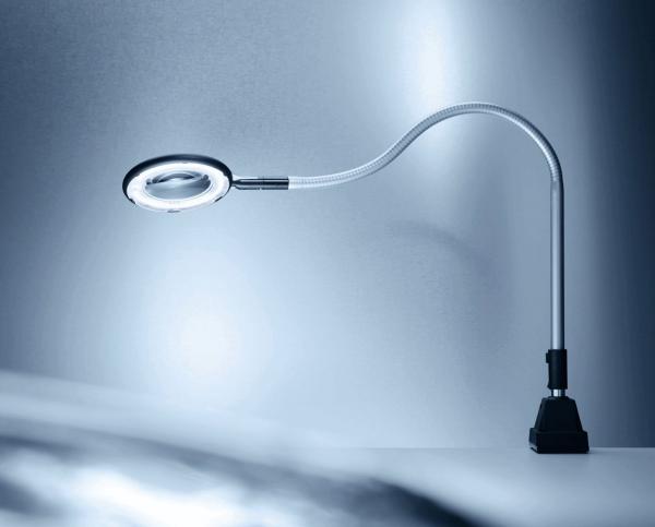 The image shows a flexible desk lamp with a round, magnifying light. The neck of the lamp is bendable and it stands on a stable, black base. The background is in soft shades of gray.