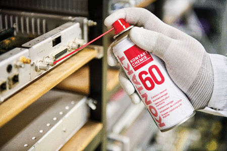 Eine Hand mit einem weißen Handschuh hält eine Spraydose. Der Sprühkopf richtet sich auf einen elektronischen Gegenstand in einem Regal. Die Dose hat die Aufschrift ?KONTAX 60?.