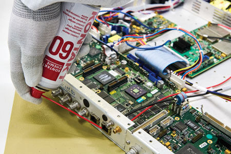 Eine Hand mit einem roten Sprühkleber hält die Dose über eine elektronische Platine. Auf der Platine sind viele farbige Drähte, Chips und Bauteile sichtbar.