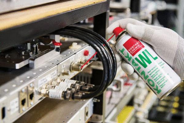Eine Hand in weißem Handschuh sprüht ein Produkt aus einer roten Spraydose auf eine technische Gerätefläche mit Kabeln und Anschlüssen. Der Hintergrund ist verschwommen.