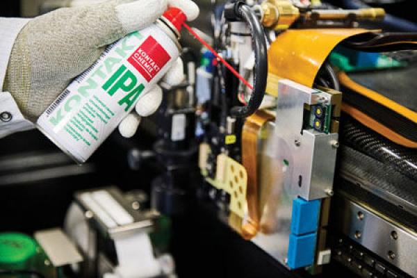 Eine Hand mit einem Sprühbehälter, der Reinigungsmittel ausstößt, hält den Behälter über eine mechanische Vorrichtung. Teile in verschiedenen Farben sind sichtbar, darunter Metall und Kunststoff.