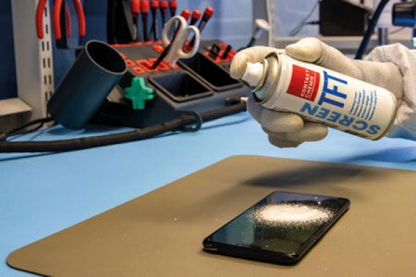 On a table, there is a smartphone. Someone is holding a spray can labeled "SCREEN TFT" and is spraying it. In the background, tools are visible.