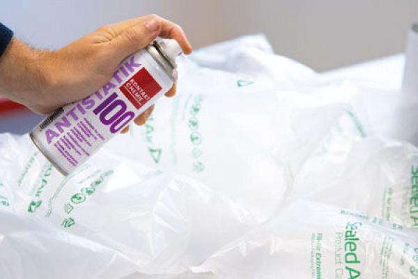 Eine Hand hält eine Spraydose mit der Aufschrift "ANTISTATIK 100". Im Hintergrund liegen aufgereihte, aufblasbare Verpackungen, die zum Schutz von Gegenständen dienen.