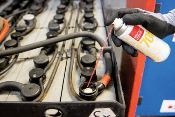 Eine Hand trägt einen schwarzen Handschuh und sprüht ein Produkt aus einer Dose auf die Kontakte einer großen, rechteckigen Batterie mit vielen Kabeln. Der Hintergrund ist blau.