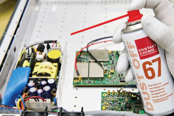 A hand wearing white gloves holds a spray can with a red nozzle. Inside an open electronic device, circuit boards and cables are visible, surrounded by various components.