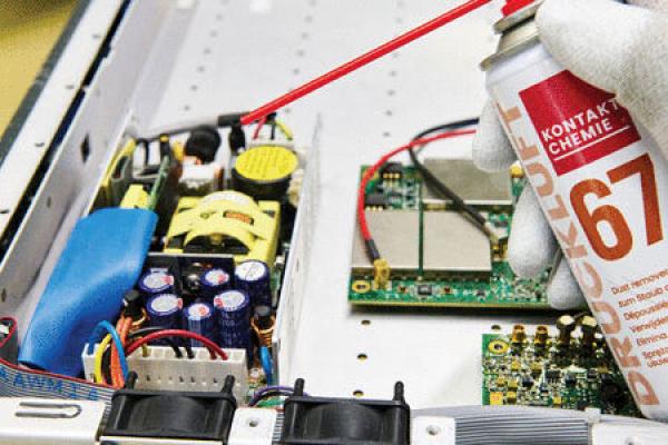 Das Bild zeigt eine Hand mit einem Spray in einem weißen Handschuh, die Kontaktreiniger auf eine elektronische Baugruppe sprüht. Es sind verschiedene farbige Bauteile innerhalb eines Geräts sichtbar.
