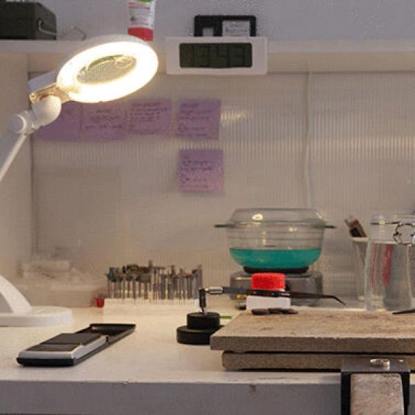 A workspace with a light magnifier, a digital display, tools in a container, a bowl with green contents, and notes on the wall. Everything neatly arranged.