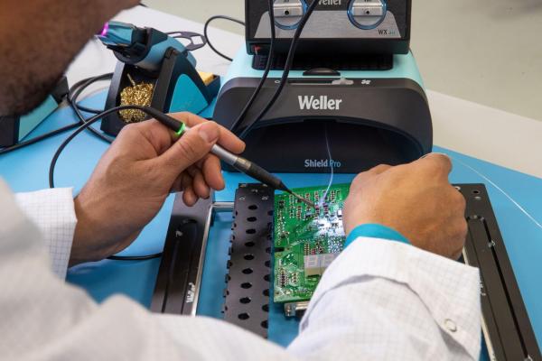 A person in a white coat is soldering with a soldering iron on a green circuit board. In the background, a Weller device is visible. The table is light blue.