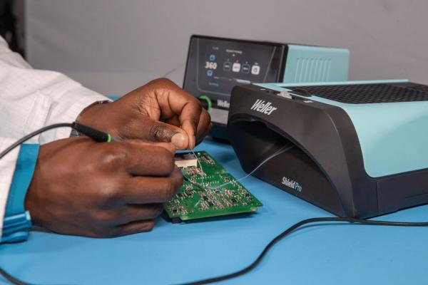 A hand works on a green circuit board, surrounded by technical devices. A device in light blue stands next to it, used for maintenance or testing.