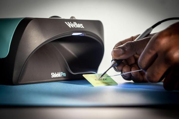A device called Weller ShieldPro is placed on a blue background. A hand is holding a card while a tool is moved over it. The light from the device shines on the card.