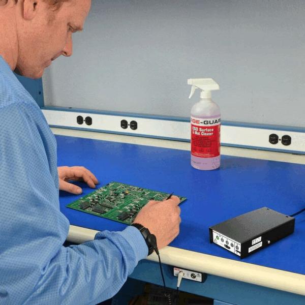 Ein Mann in einem blauen Laborkittel arbeitet an einer grünen Platine auf einem blauen Tisch. Neben ihm steht eine Sprühflasche und ein schwarzes Gerät mit Anschlüssen.