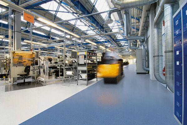 The image shows a modern factory hall with high ceilings, blue steel beams, and a bright floor. On the left side are machines and shelves, while a forklift is on the move.