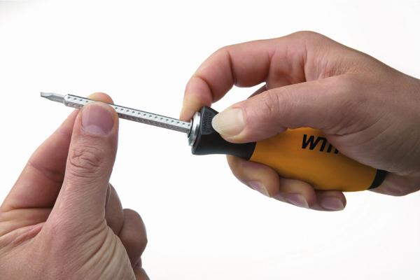 Auf dem Bild ist eine Hand zu sehen, die einen Schraubendreher mit gelbem Griff hält. Die Spitze des Schraubendrehers zeigt nach oben, während eine zweite Hand ihn vorsichtig festhält.