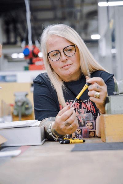 A smiling woman with long, blonde hair and glasses holds a yellow pencil. She is sitting at a table with tools and plastic-like objects, surrounded by a workshop atmosphere.
