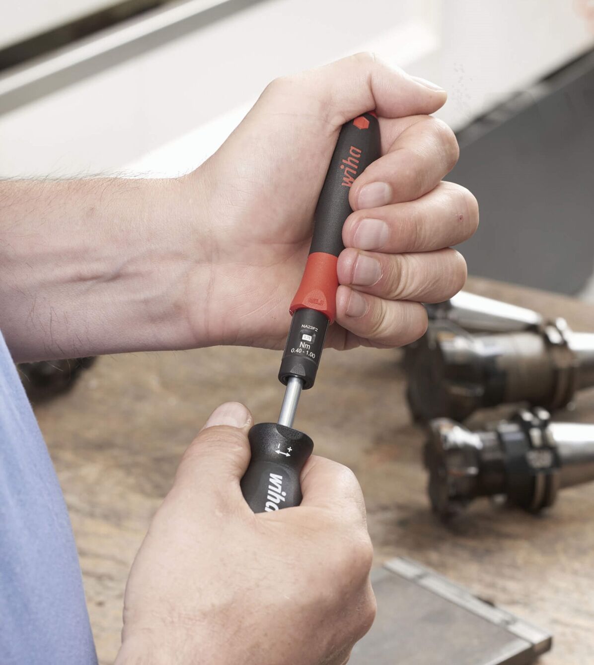 A hand holds a screwdriver with a red handle. The tool is used for working on metal parts that are lying on a table. The background is blurry.