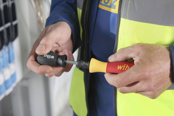 Two hands holding different screwdrivers: one black with "Wera" and one red-yellow with "Wit". The person is wearing a yellow safety vest over a blue shirt.