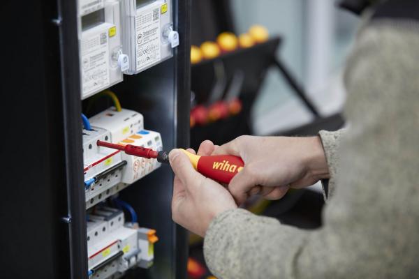 A person is holding a screwdriver and working on an electrical panel, which is equipped with multiple devices and colorful cables.