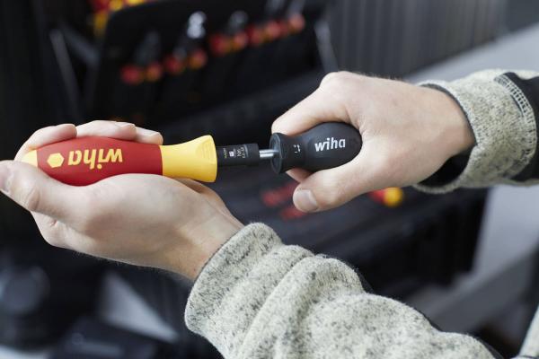 A hand holds a screwdriver with a red and yellow handle. Another small attachment is pushed onto the end. Tools are visible in the background.