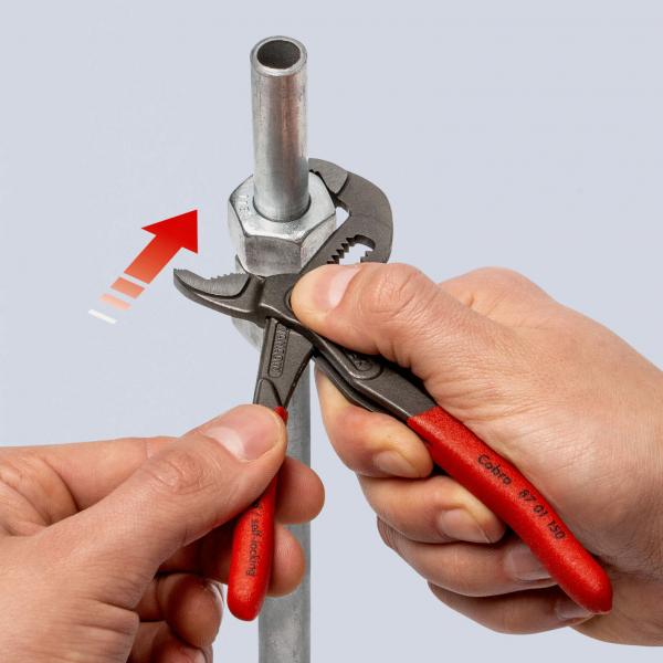 The image shows two hands, each holding a tool. One hand is using an adjustable wrench, while the other is holding a metal pipe with a nut.