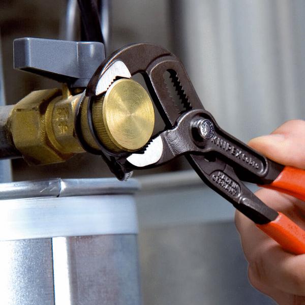 Ein Handwerker benutzt eine verstellbare Zange, um eine runde, goldene Schraube an einem silbernen Metallzylinder zu befestigen. Der Fokus liegt auf den Werkzeug und der Schraube.