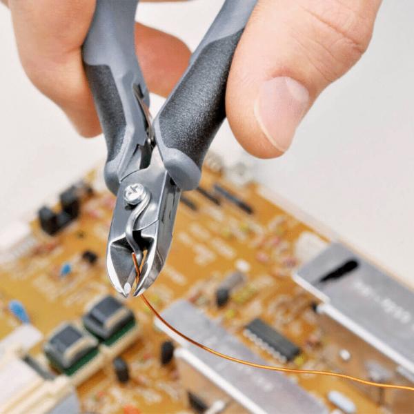 A hand is holding pliers that are cutting thin copper wire. In the background, there is a circuit board with various electronic components and a golden surface.