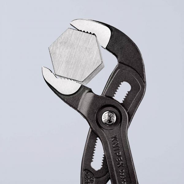 Das Bild zeigt eine schwarze Zange, die eine hexagonale Metallmutter hält. Der Hintergrund ist hell und neutral. Die Zange hat eine geschwungene Form mit rutschfesten Griffen.
