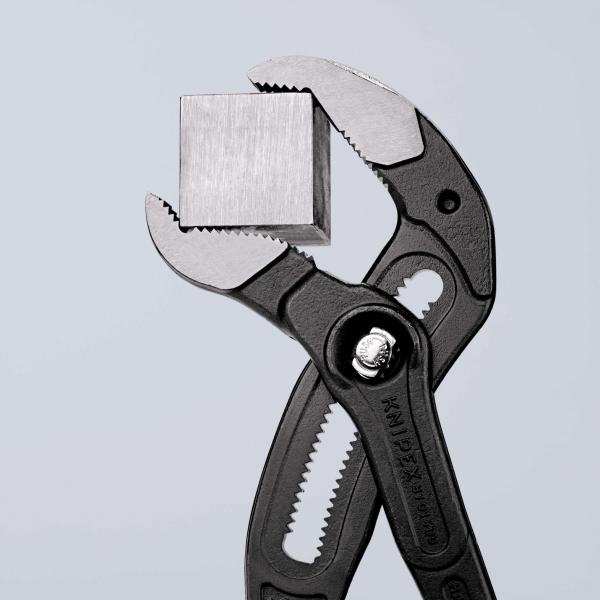 The image shows a black pair of pliers holding a small, square metal block. The background is bright and neutral.