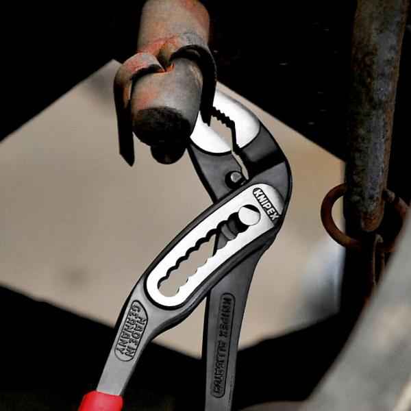 An adjustable wrench grips a thick, metallic bolt attached to an old pipe. The wrench is red and silver in color with a special clamping design.